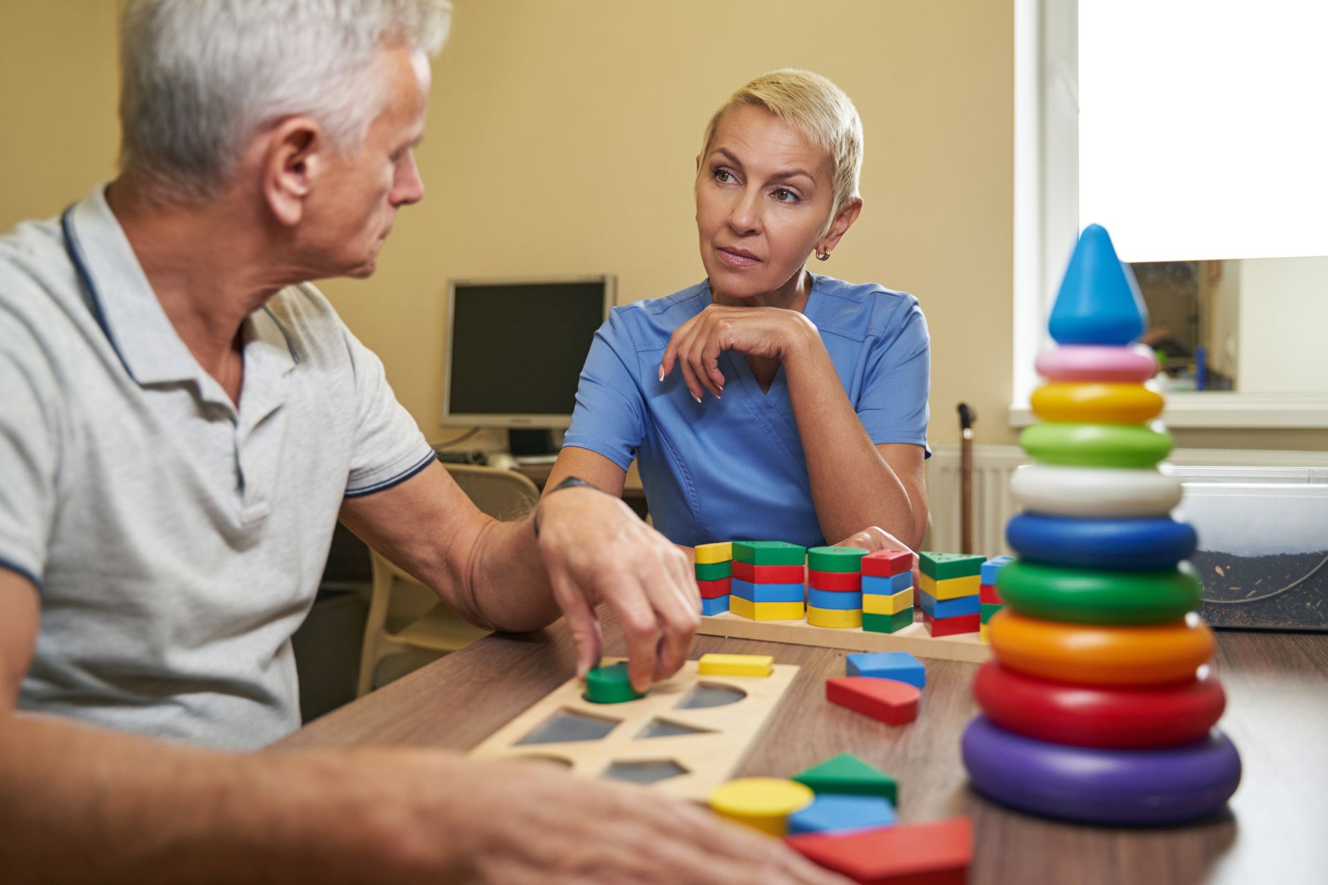 Blonde nurse with a pixie cut, wearing light blue scrubs, sitting next to an elderly man with gray hair, dressed in a white shirt, while engaging in toy figure therapy for cognitive stimulation.