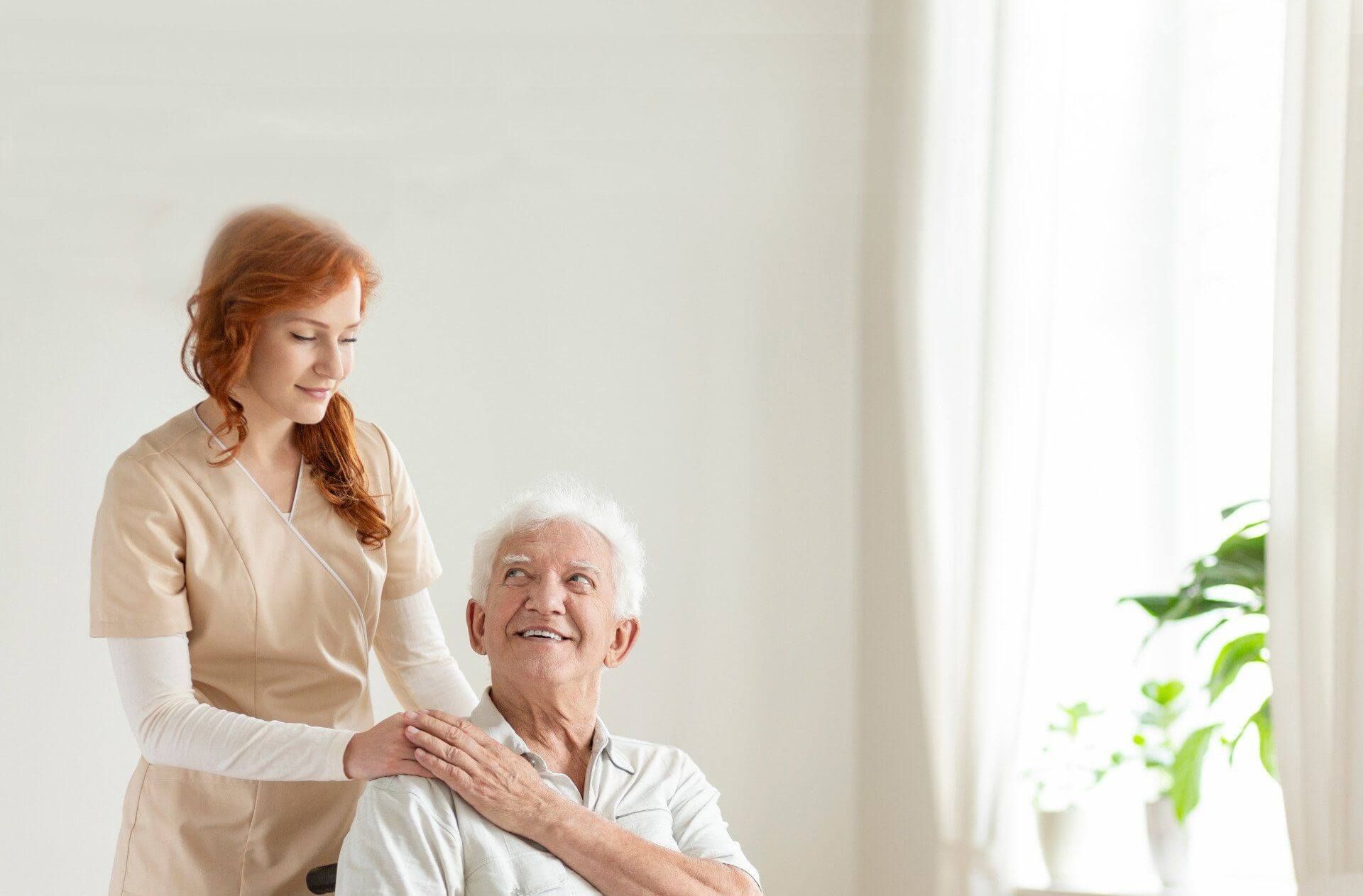 Red-haired nurse in beige scrub gently touching the shoulder of an elderly man with gray hair and eyebrows, smiling up at her, seated in a wheelchair.