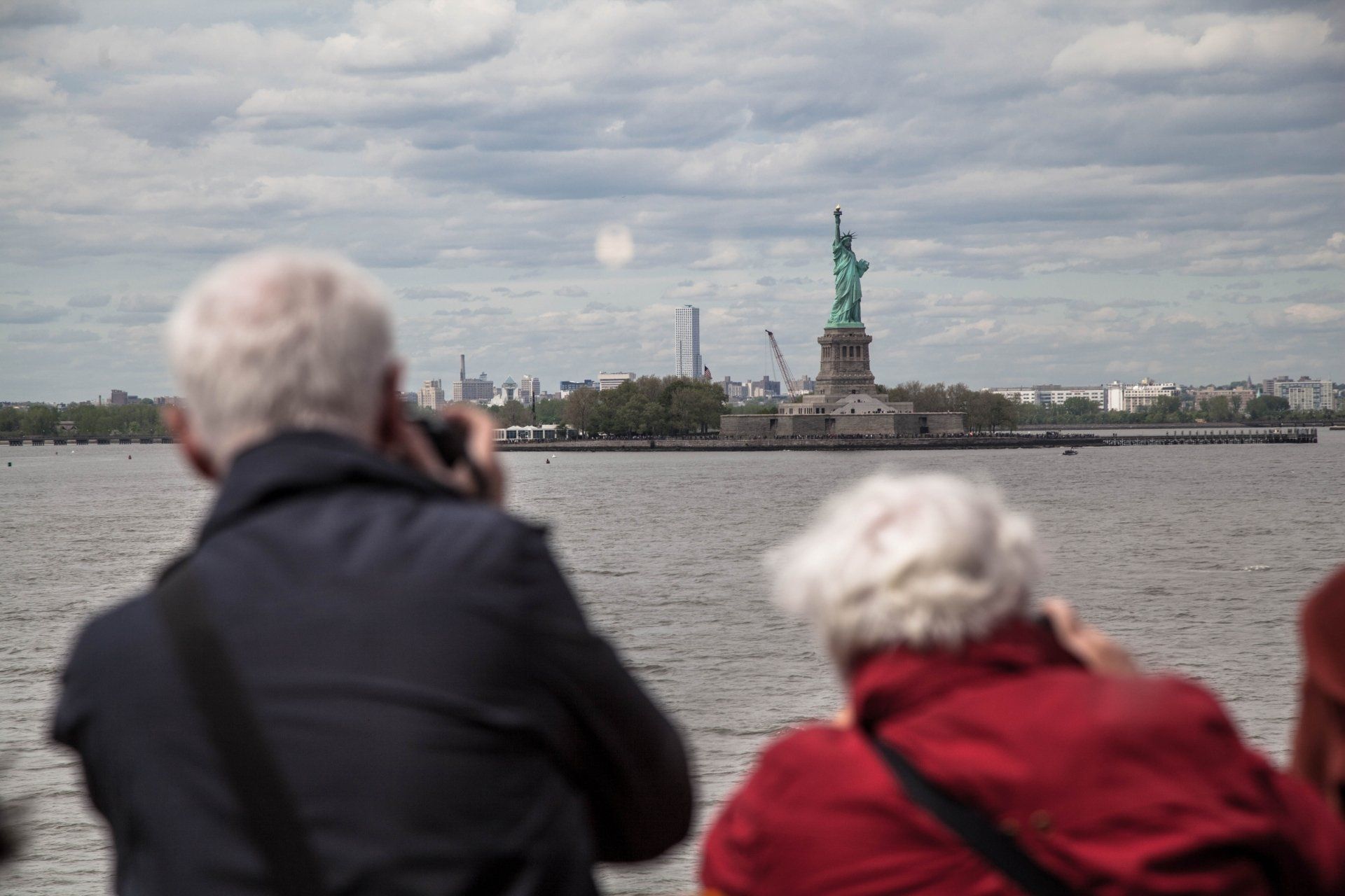 Elderly couple with gray hair, back-facing, wearing coats, on a ferry, taking photos of the Statue of Liberty.