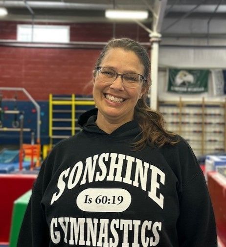 A woman wearing a sonshine gymnastics hoodie is smiling in a gym.