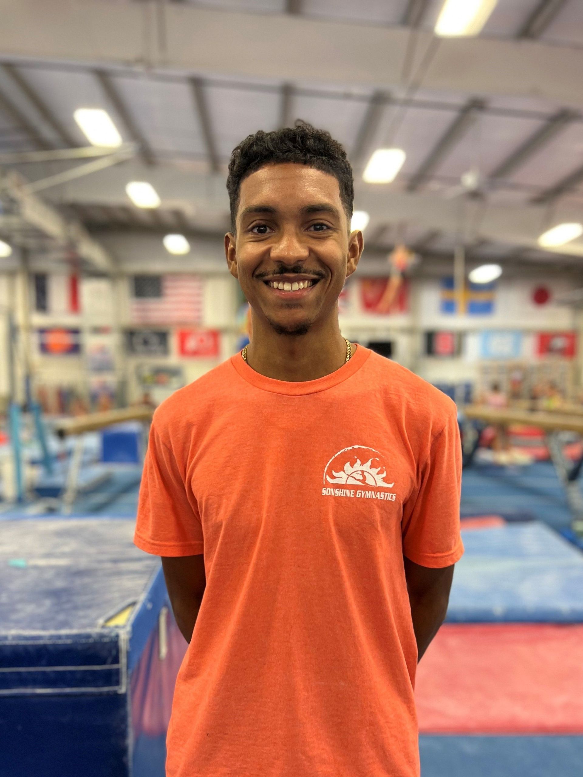 A young man in an orange t-shirt is smiling in a gym.