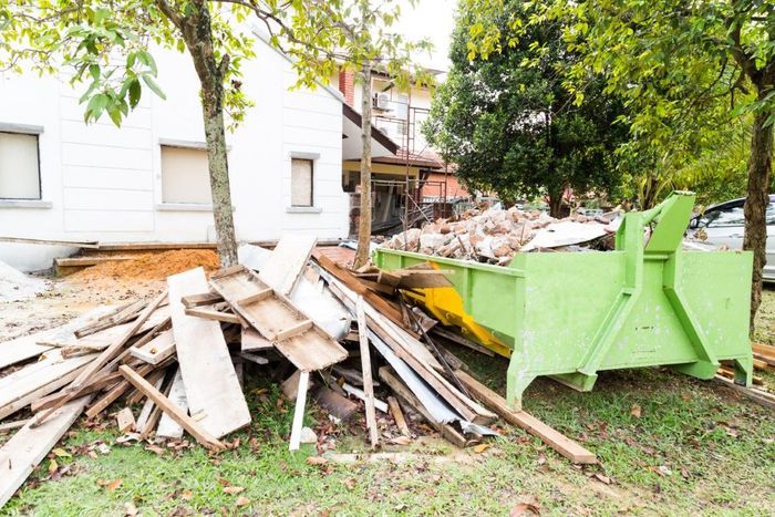 A green dumpster filled with debris from a building renovation. Wood and other materials are piled nearby on a lawn, with a white house in the background.