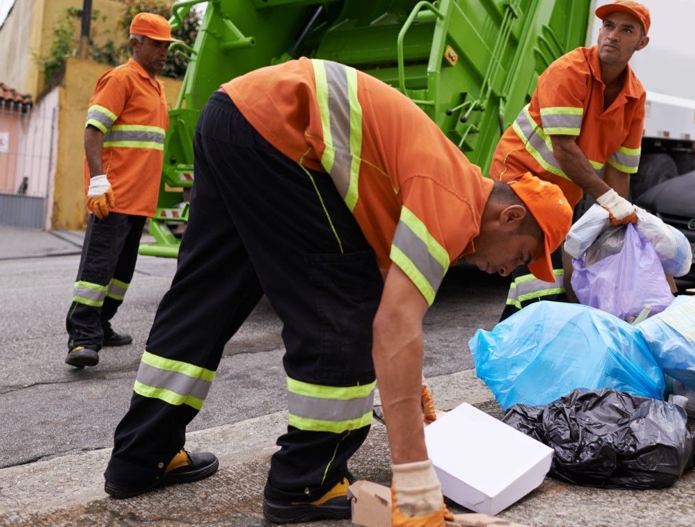 Three sanitation workers in orange uniforms load trash bags into a green garbage truck on a city street.