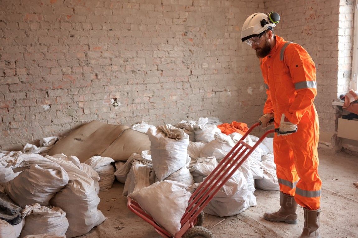 Construction worker in orange jumpsuit uses a hand truck to move white bags in a room with exposed brick walls.