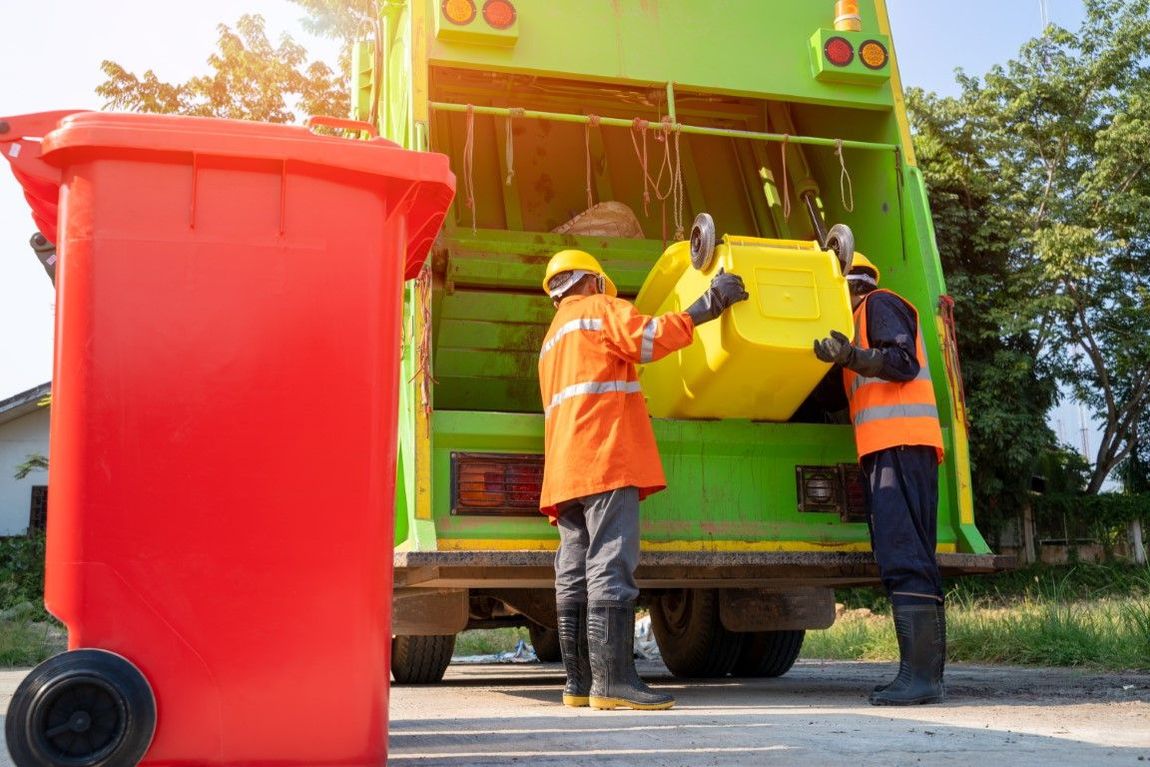 Two sanitation workers loading yellow bin into a green garbage truck, red bin in foreground.