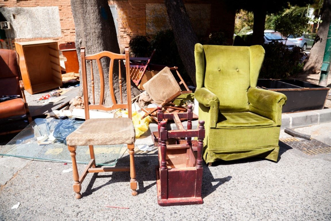 Discarded furniture on a city street: a wooden chair, green armchair, and small red box.