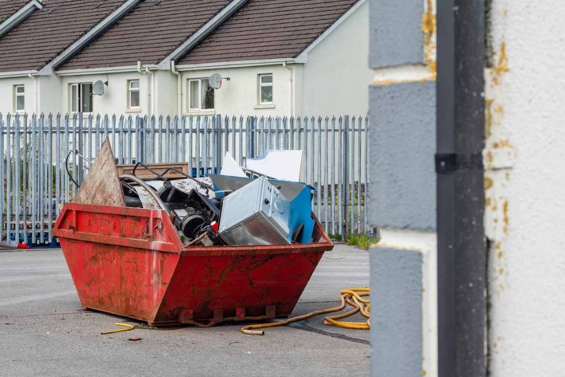 Red dumpster overflowing with metal and construction debris, set against a background of houses and a metal fence.