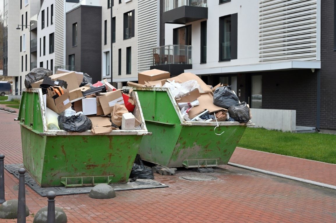 Two overflowing green dumpsters on a brick walkway in front of a modern apartment building.