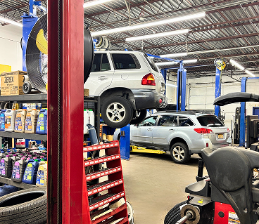 Inside a car repair shop, a silver SUV is raised on a lift above a smaller silver car, with shelves of oil in the foreground. | Smithwest Service Center