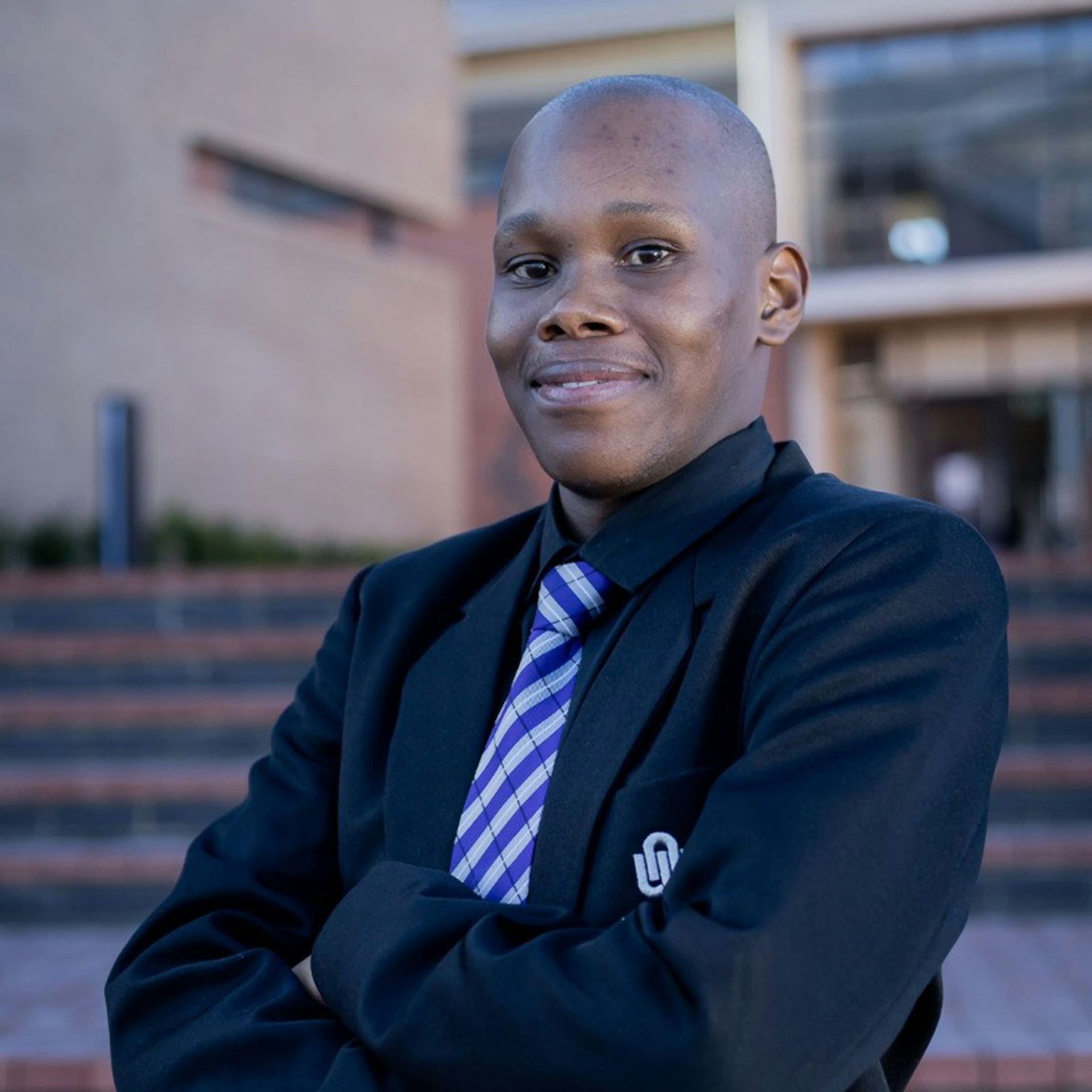 Man with shaved head in a blazer and tie, arms crossed, smiling in front of a building.