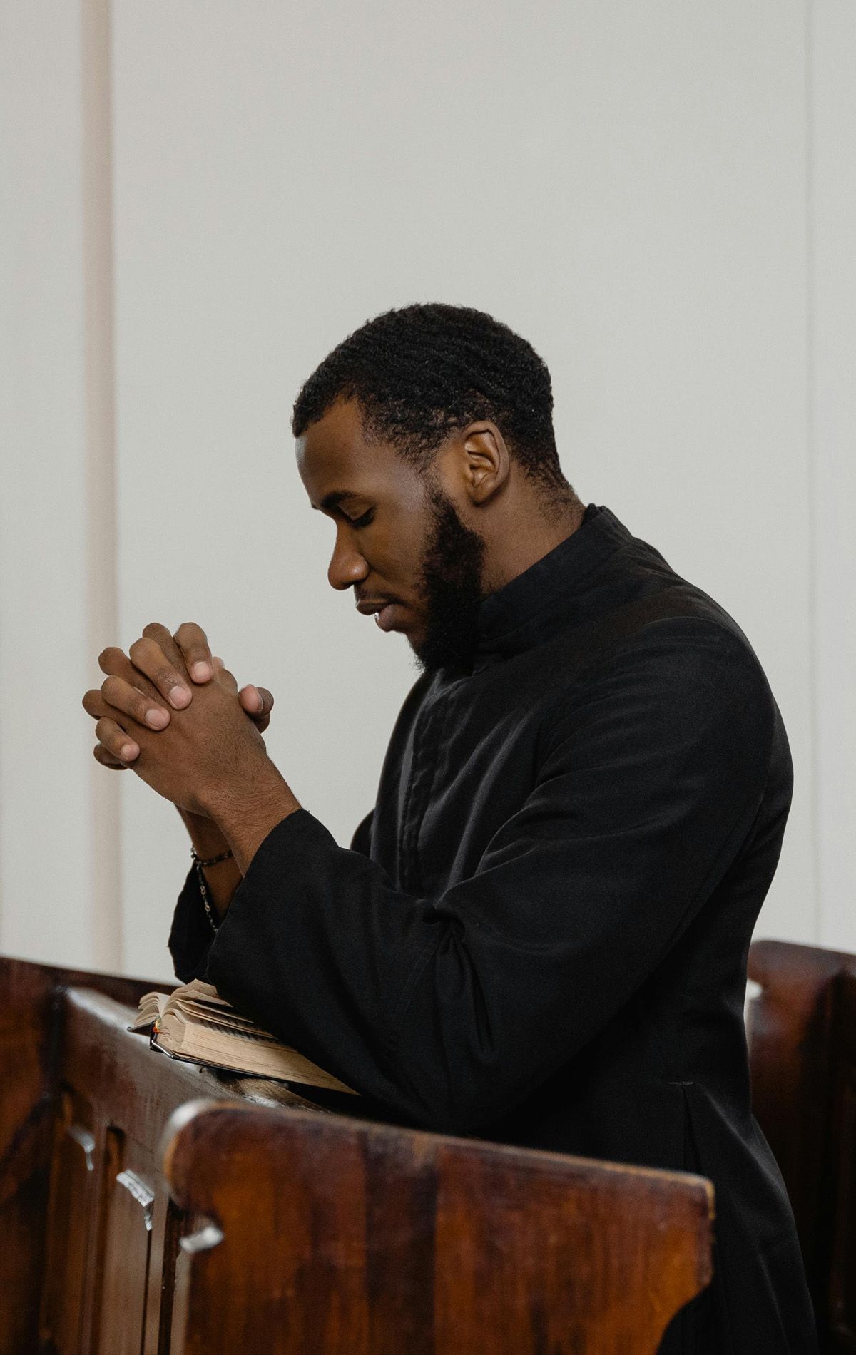 Man with folded hands in prayer, leaning on wooden pew, inside a church.