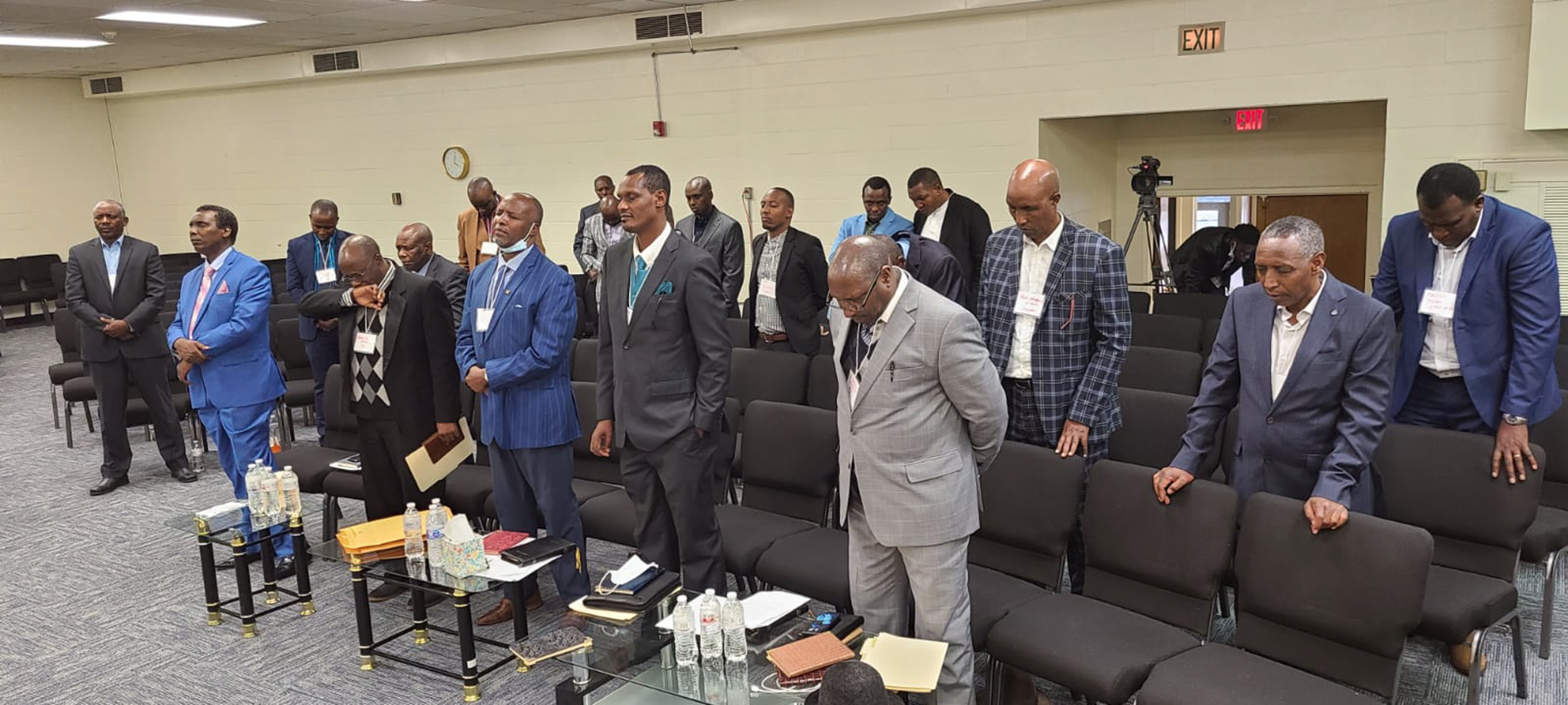 A group of formally dressed men stand and bow their heads in an auditorium setting.
