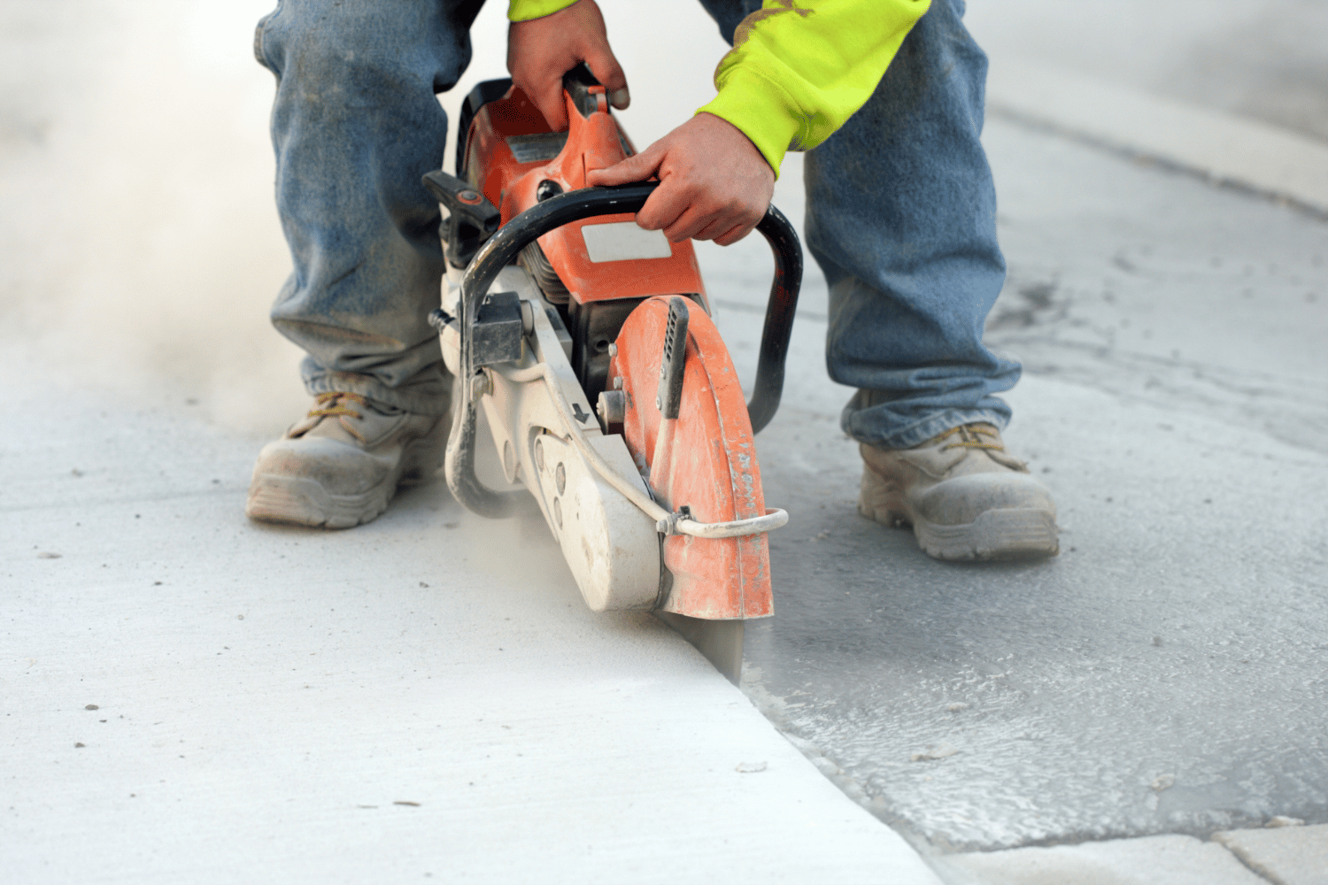 A worker uses a handheld orange power saw to cut a line into a concrete sidewalk, creating dust.