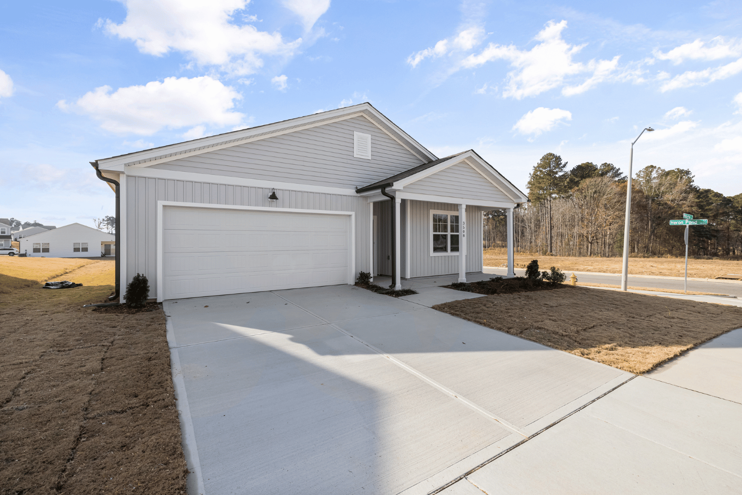 A single-story, light gray house with a two-car garage and front porch, surrounded by dry grass under a blue sky.