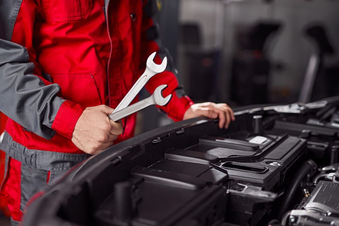 A mechanic in a red and grey uniform holds two wrenches while working on an open car engine in a garage.