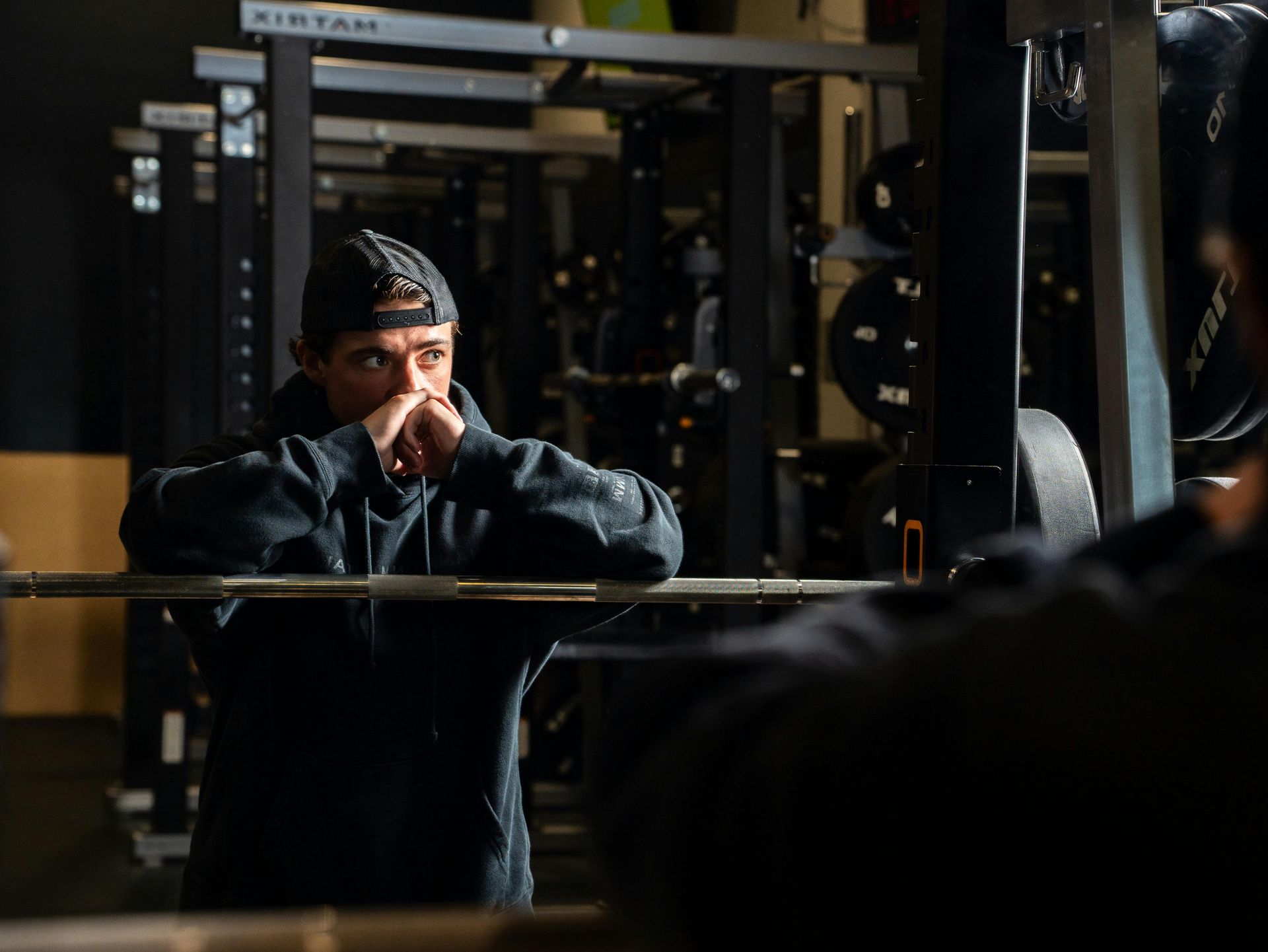 Man in a gym, leaning on barbell, contemplating, with hand on face. Rack and weights in background.