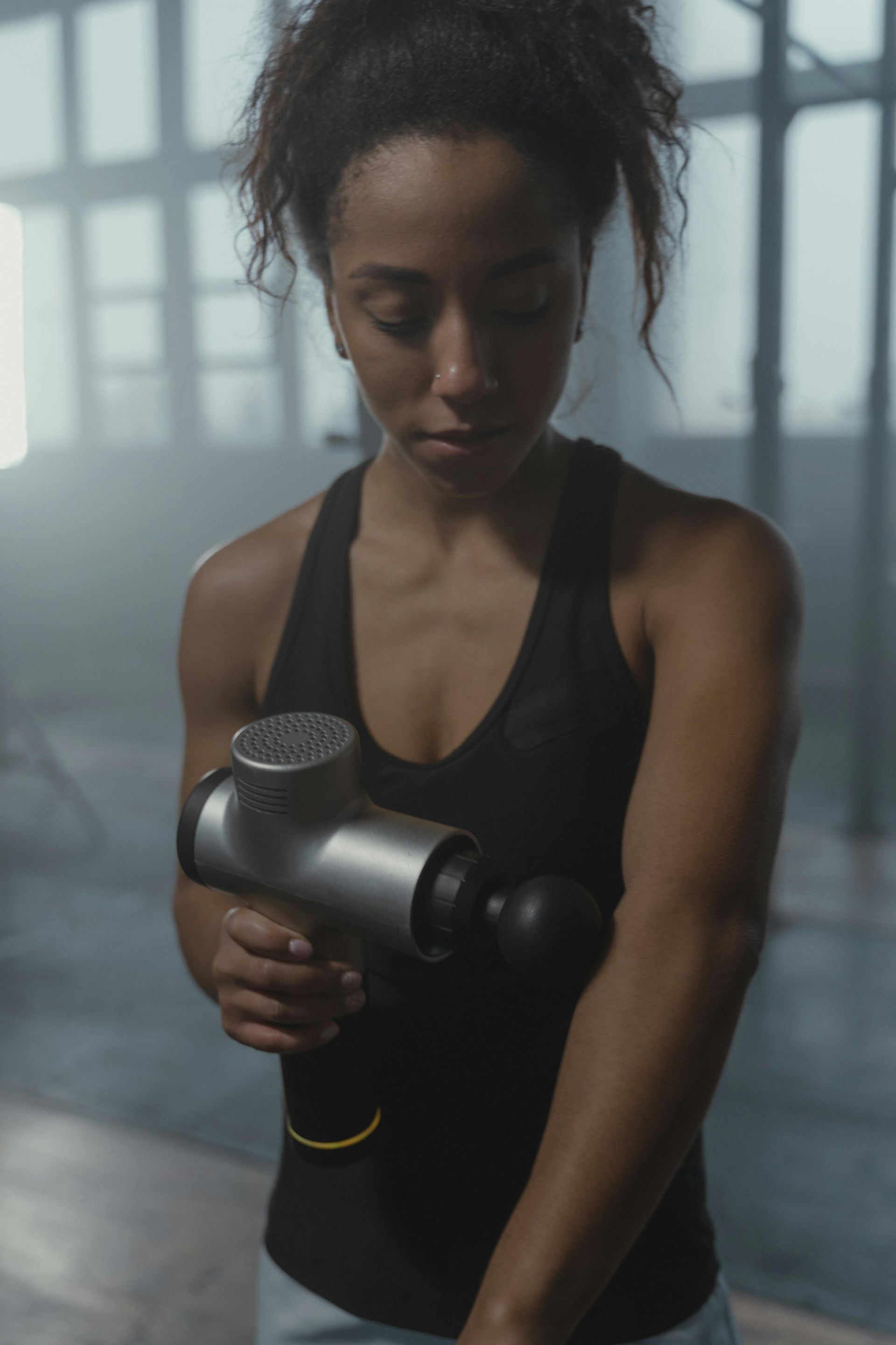 Woman in black tank top using massage gun on her arm in gym.
