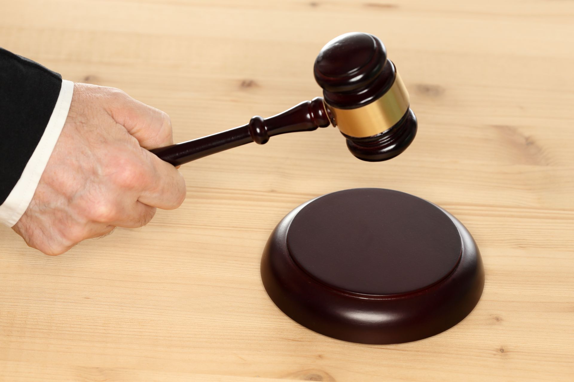 Hand holding a gavel above its wooden base, on a light wood surface.