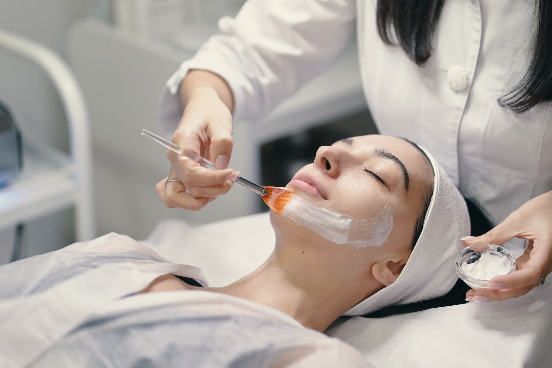 Woman receiving facial treatment, esthetician applying mask with a brush.