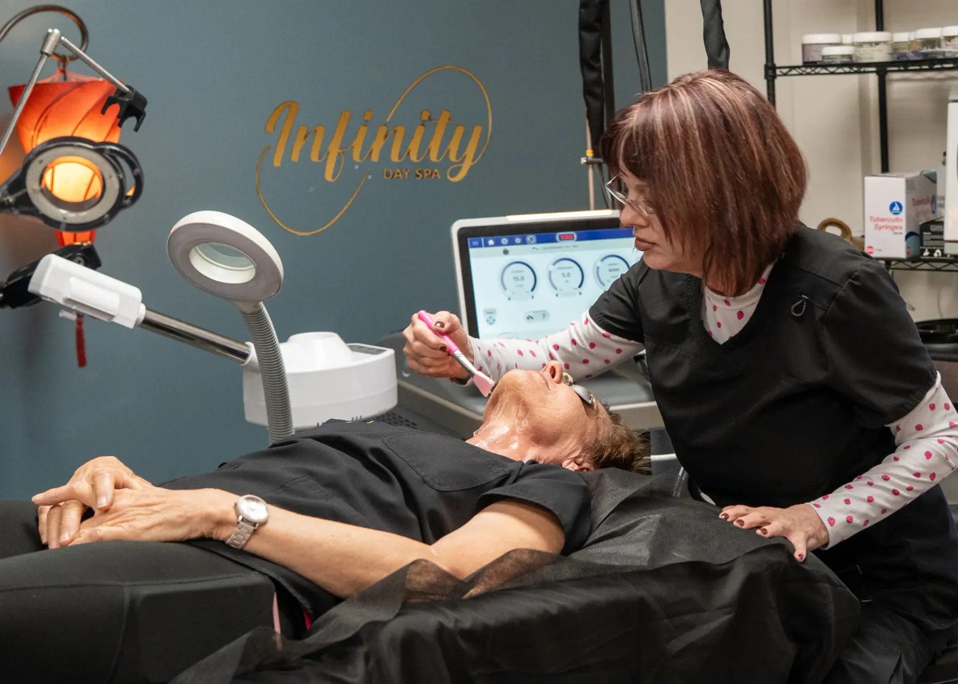 A woman receives a facial treatment from a skincare professional at Infinity. The patient lies on a treatment bed under bright lights while the technician uses a tool on her face.