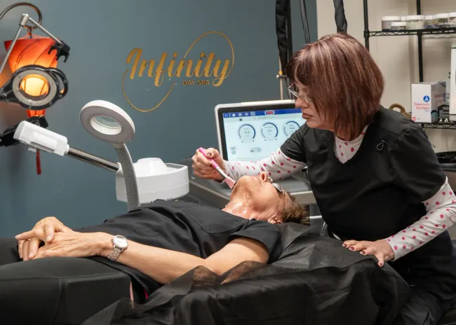 A technician performs a cosmetic procedure on a client lying down. The setting is an Infinity Aesthetics clinic.