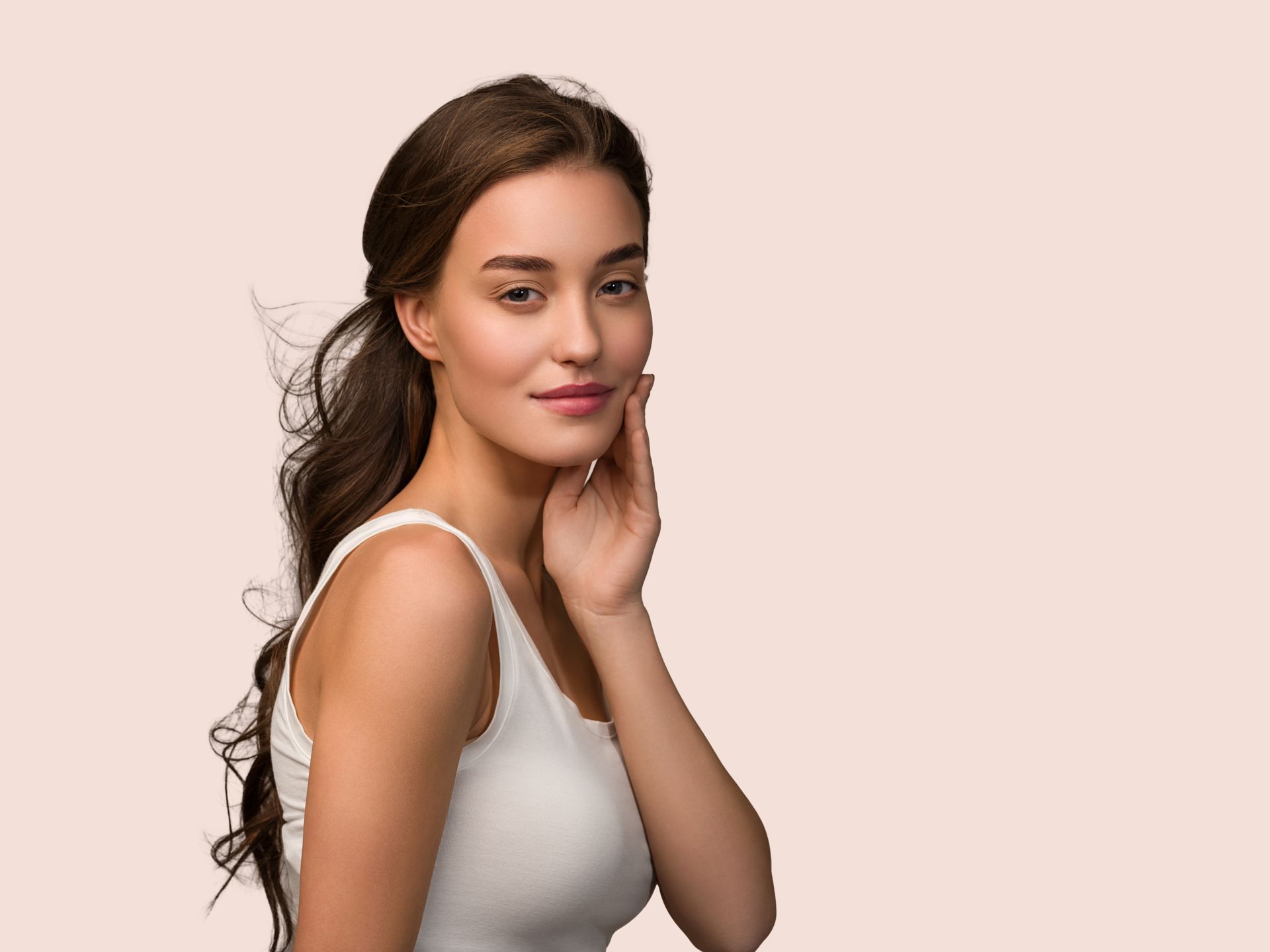 A woman in a white tank top is touching her face and smiling.