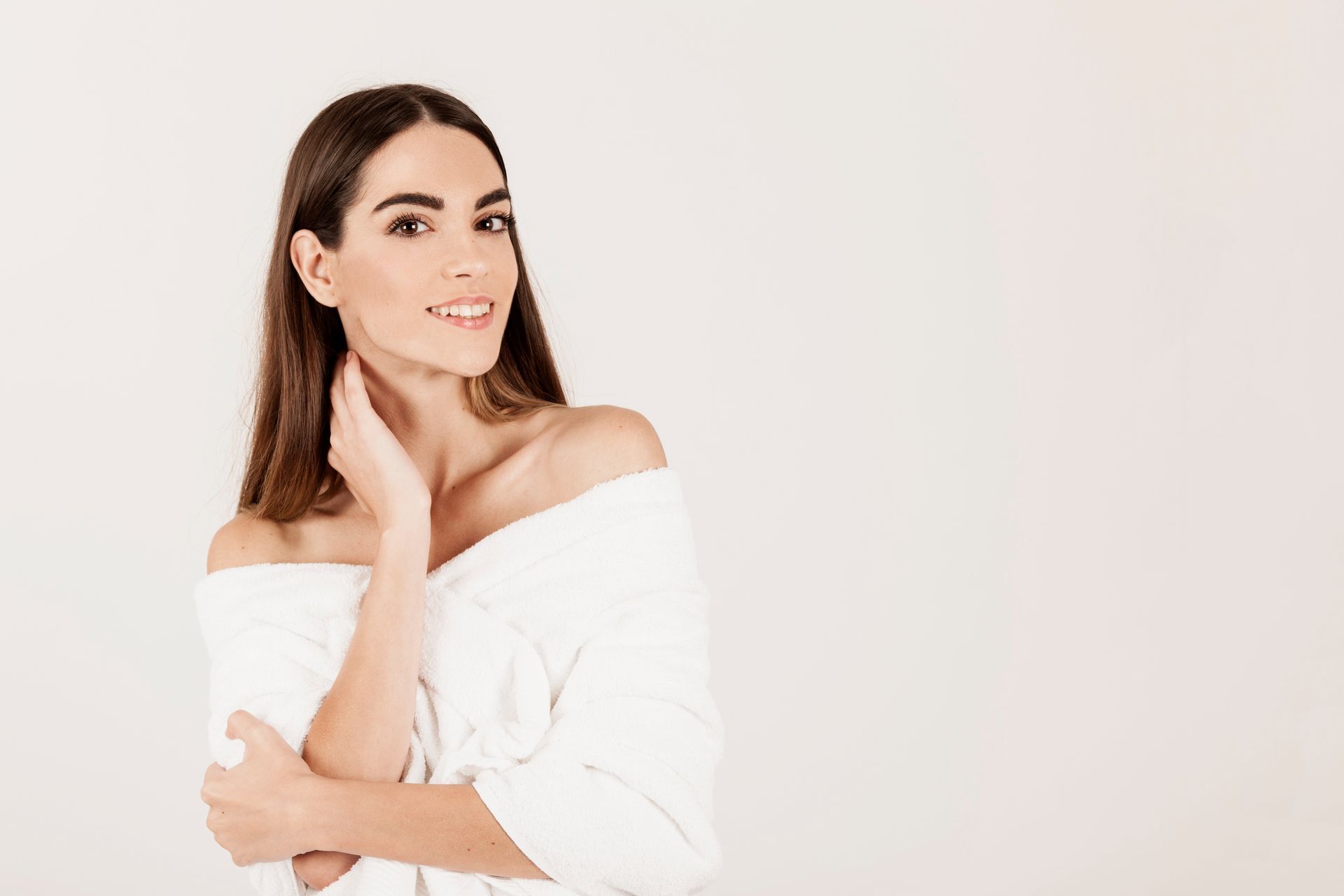 Woman with long brown hair wearing a white off-the-shoulder top, smiling and touching her neck, against a white backdrop.
