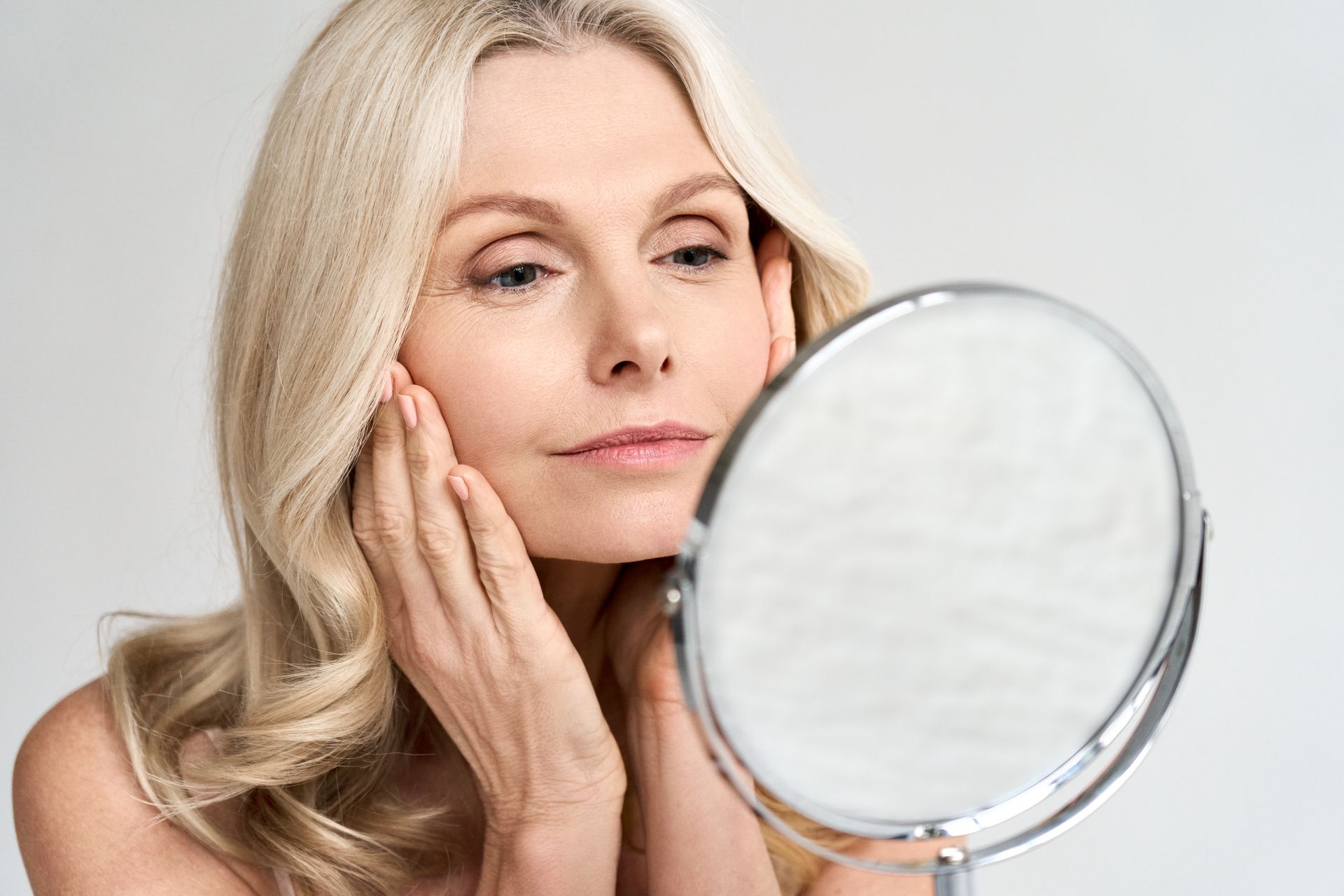 Blonde woman examining her face in a round mirror, looking concerned, indoors.