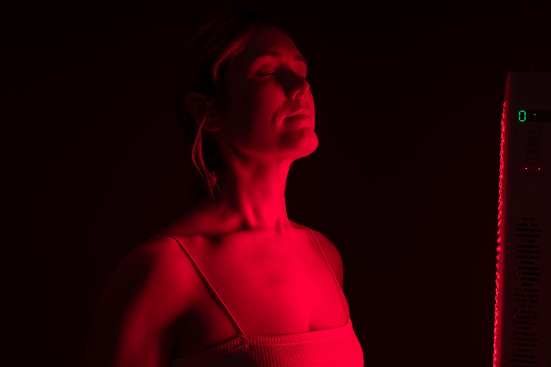 Woman illuminated by red light therapy, head tilted upward, in a dark room. The therapy device is visible.