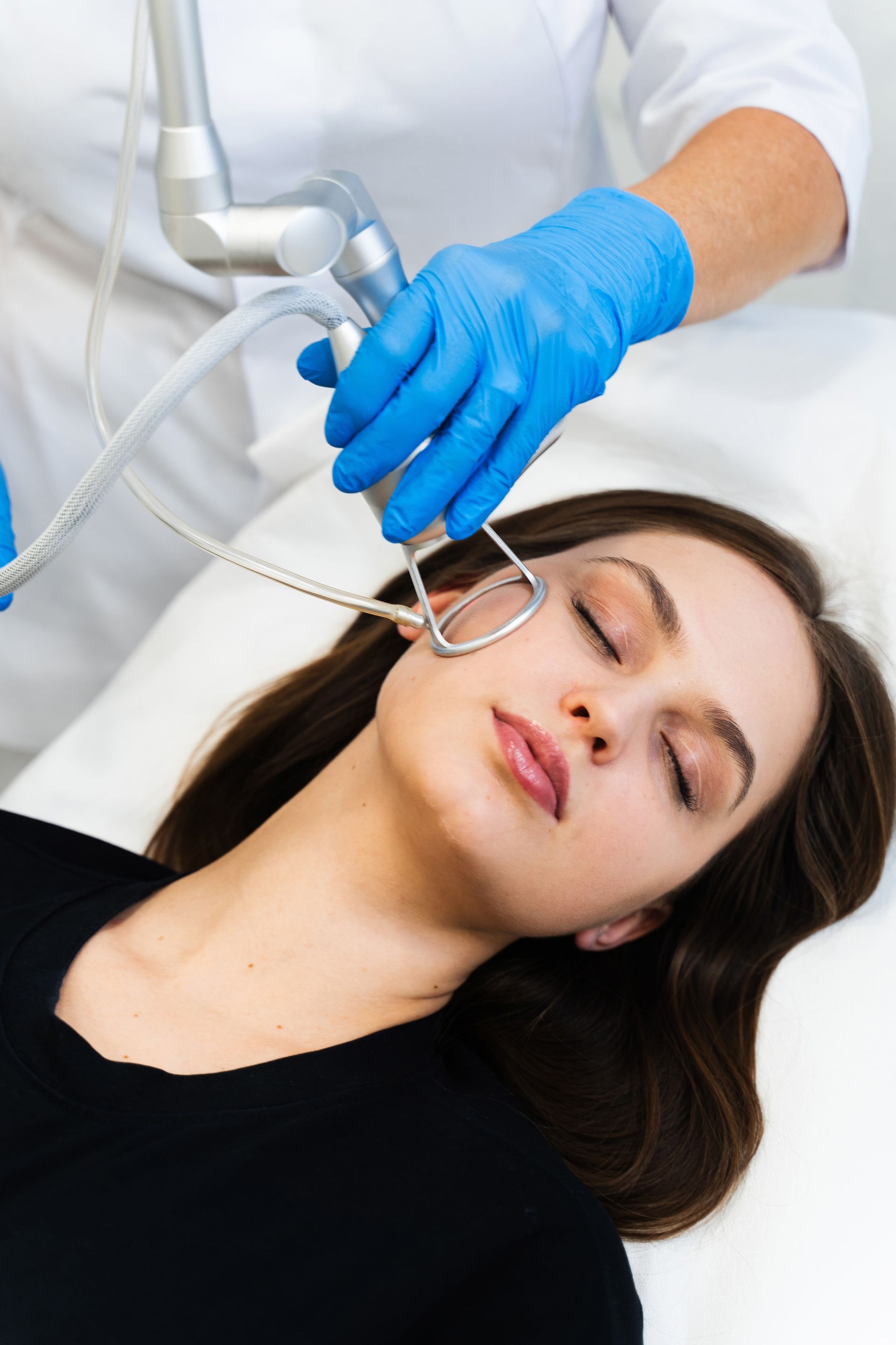 Woman receiving laser facial treatment; technician in blue gloves holds device near her face, indoors.