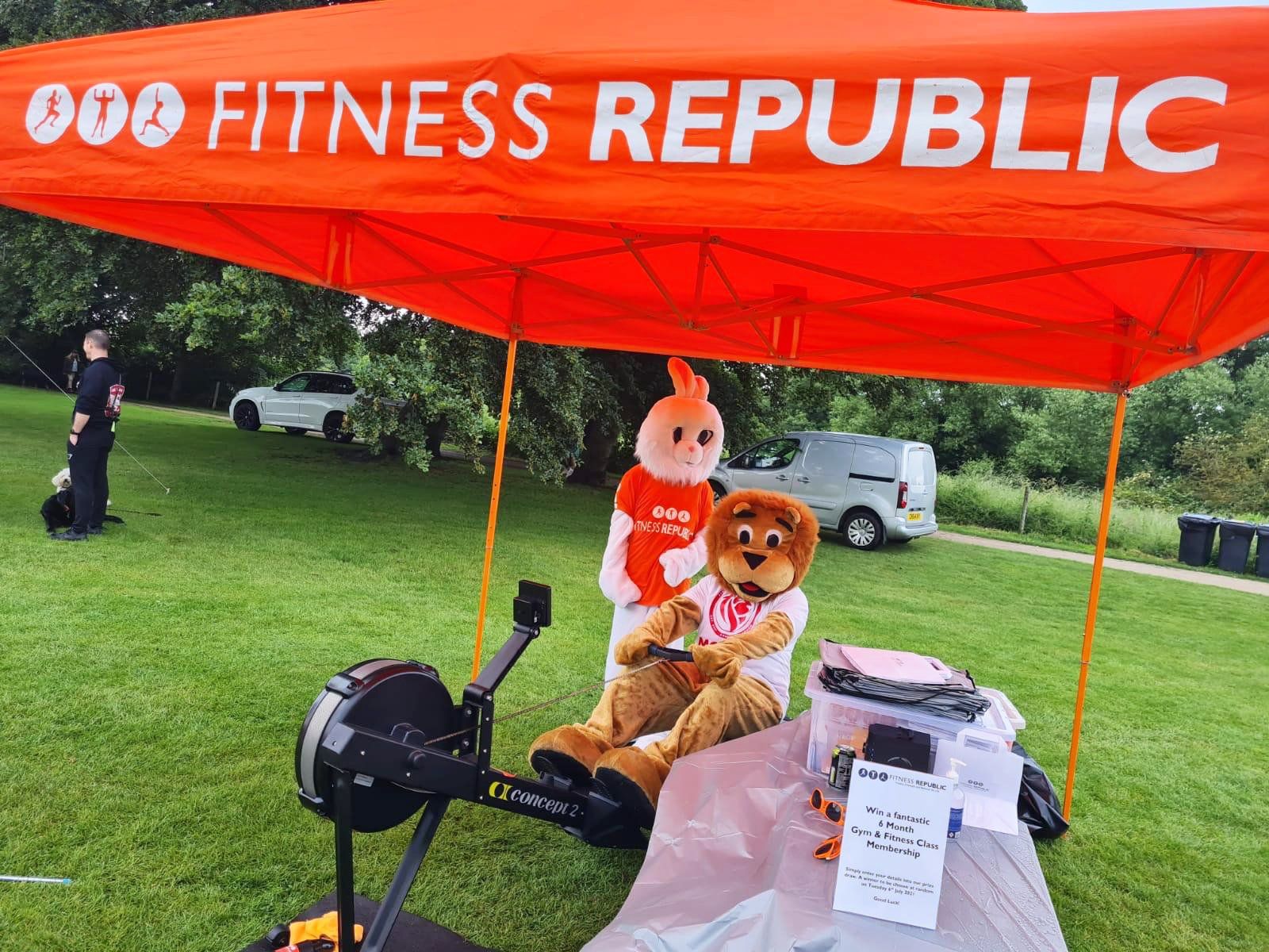 A mascot is sitting under an orange tent that says fitness republic