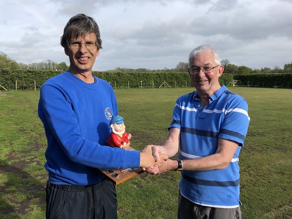 Two men are shaking hands in a field while one of them is holding a gnome.
