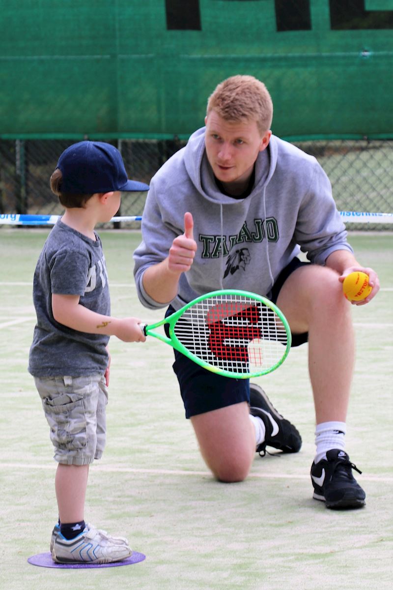 A man is kneeling down with a tennis racquet and giving a thumbs up to a little boy