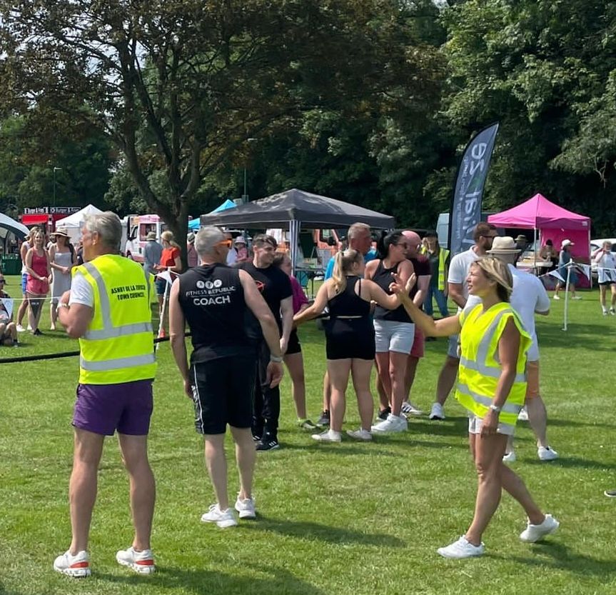 A group of people are standing in a grassy field with a man wearing a yellow vest that says ' emergency services '