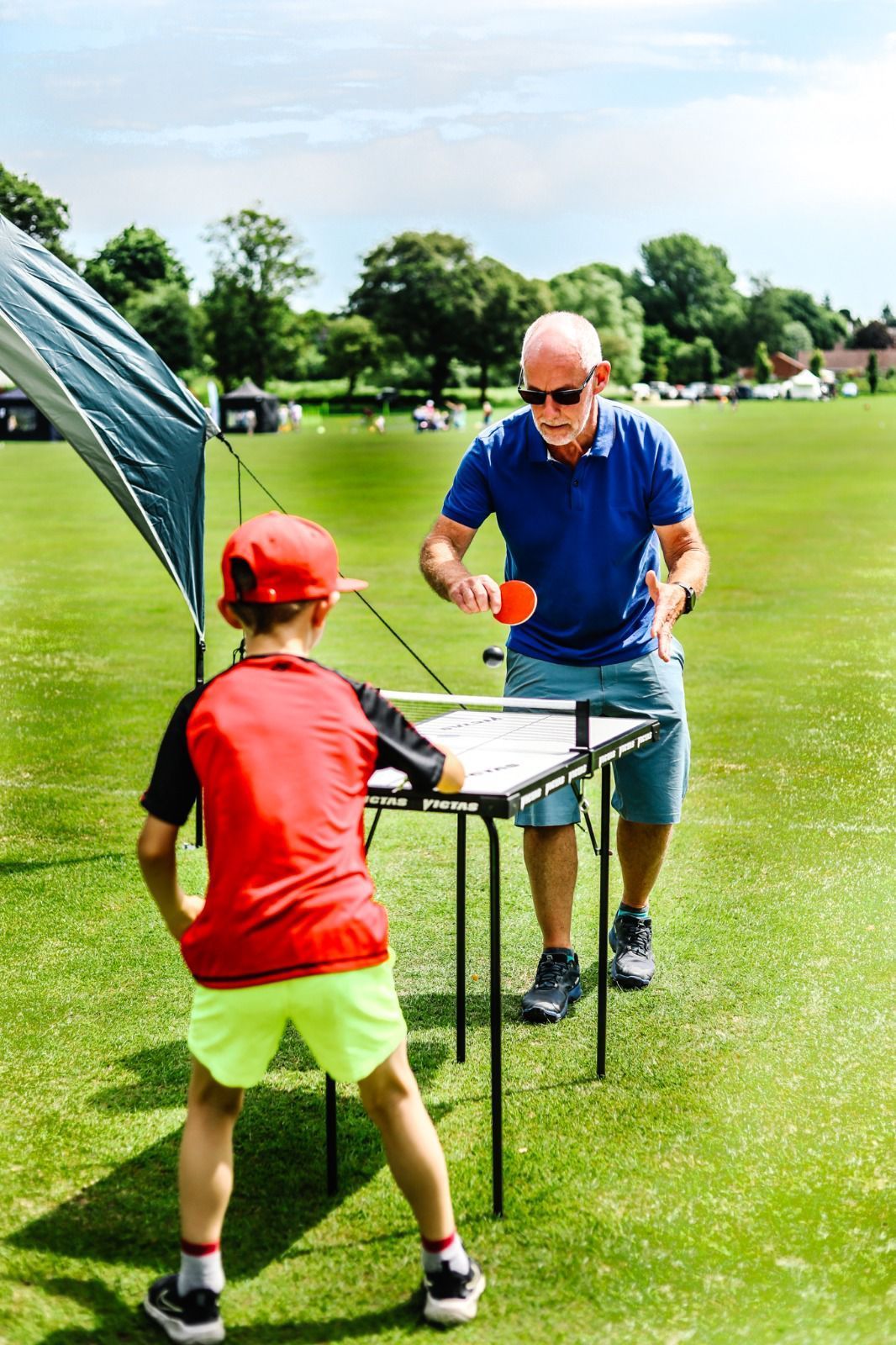 A man and a boy are playing ping pong on a table in a field.