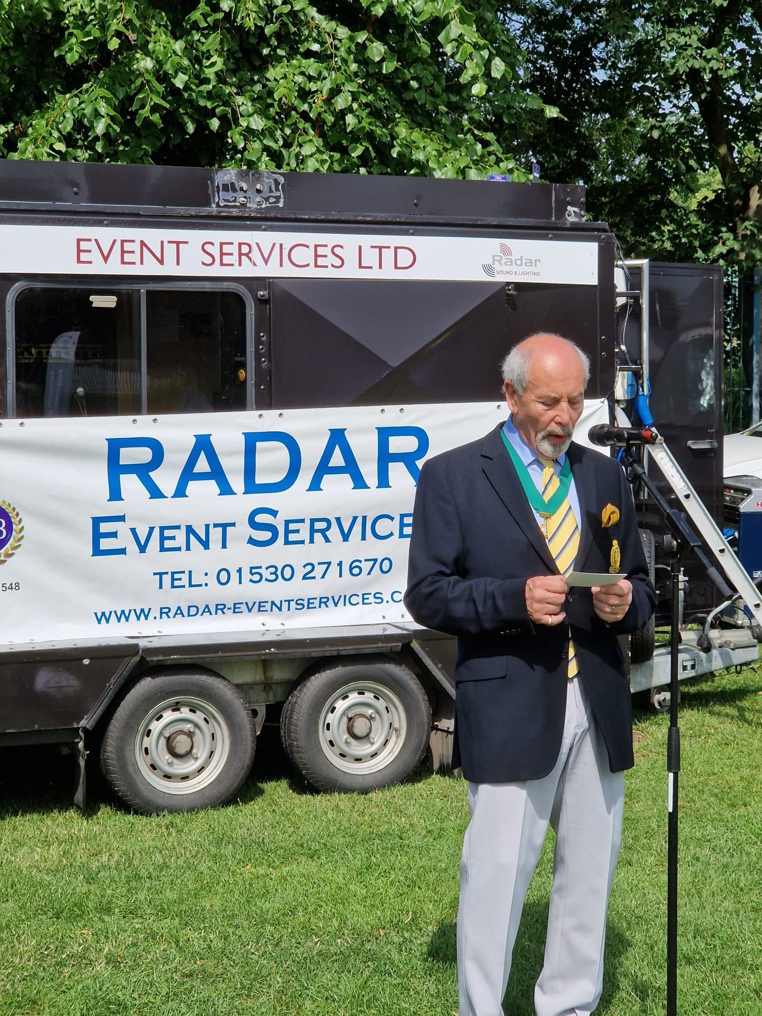 A man is standing in front of a microphone in front of a radar event service trailer.