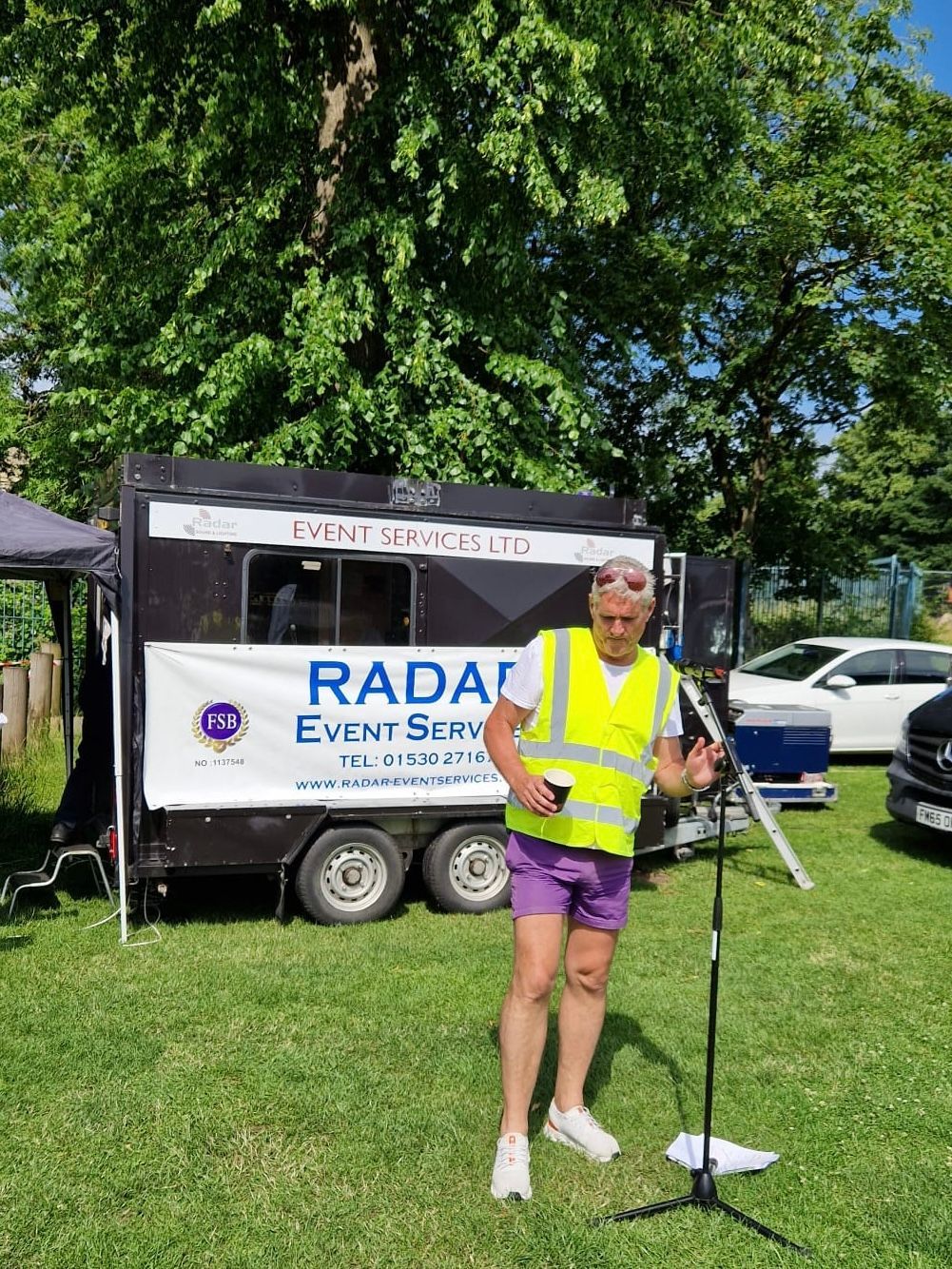 A man in a yellow vest is standing in front of a microphone in front of a trailer.