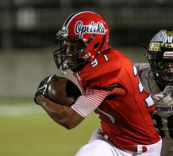 Lacrosse player in orange shirt prepares to shoot, sunlight in background.