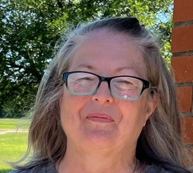 A woman wearing glasses is standing in front of a brick wall.