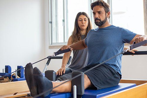 A man is sitting on a pilates machine with a woman standing behind him.
