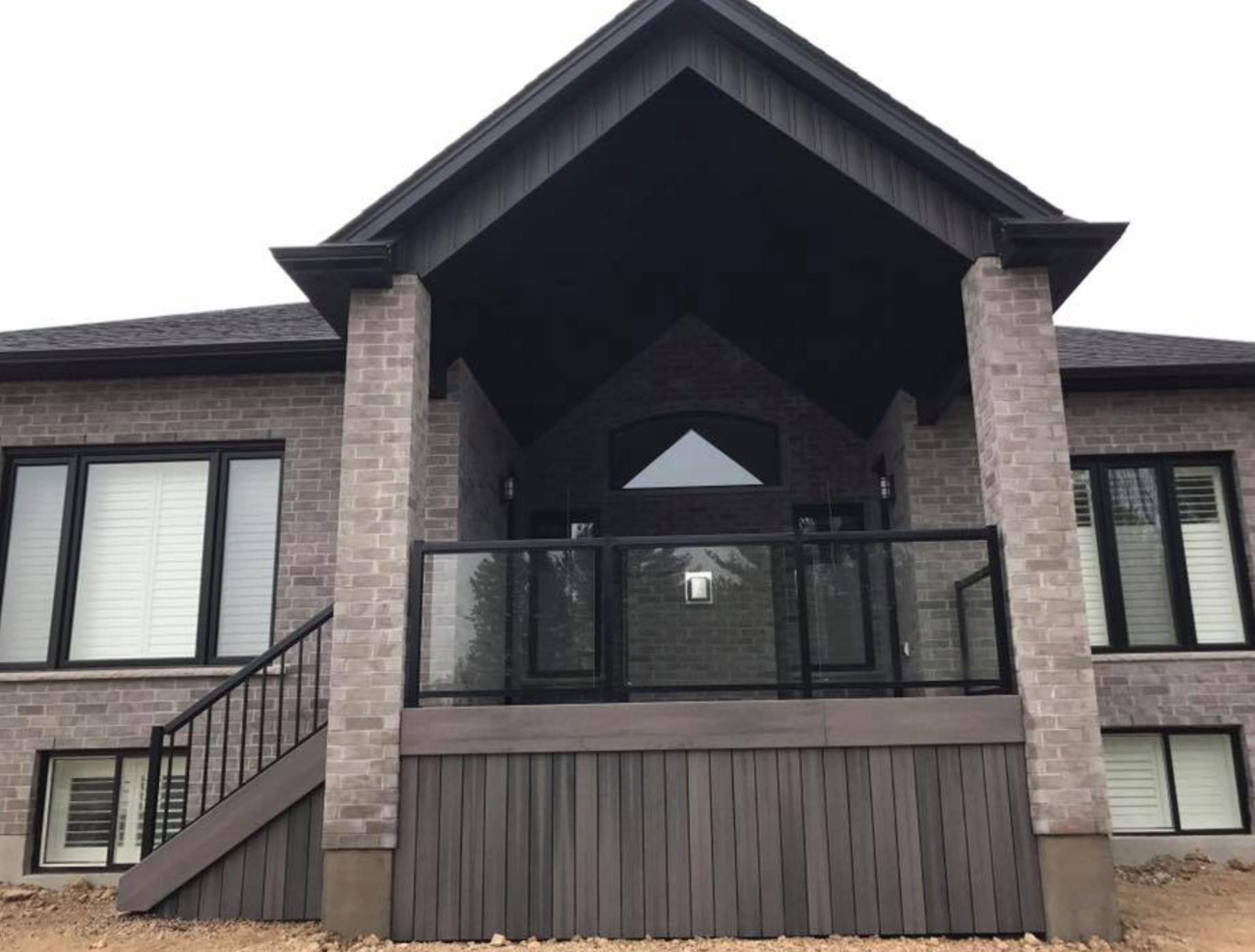 A gray brick house with a black gable roof over a glass-railed deck.