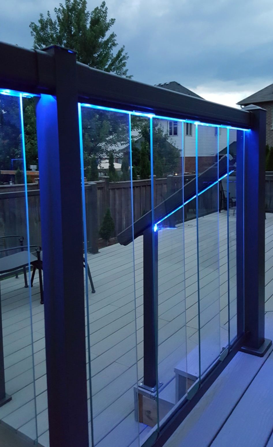 Blue-lit glass railing on a deck, dark gray frame, outdoor setting with backyard view.