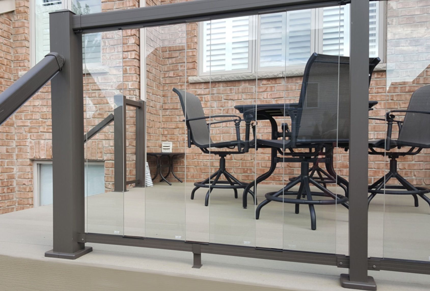 Deck with clear glass panels, dark brown frame, and outdoor furniture. Brick house in the background.