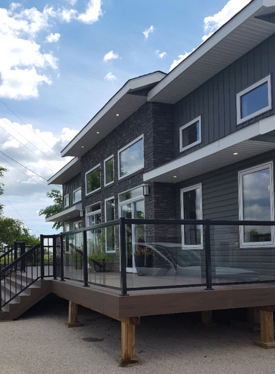 Modern two-story house with gray siding and brick facade, featuring a deck with glass railing and a cloudy sky.