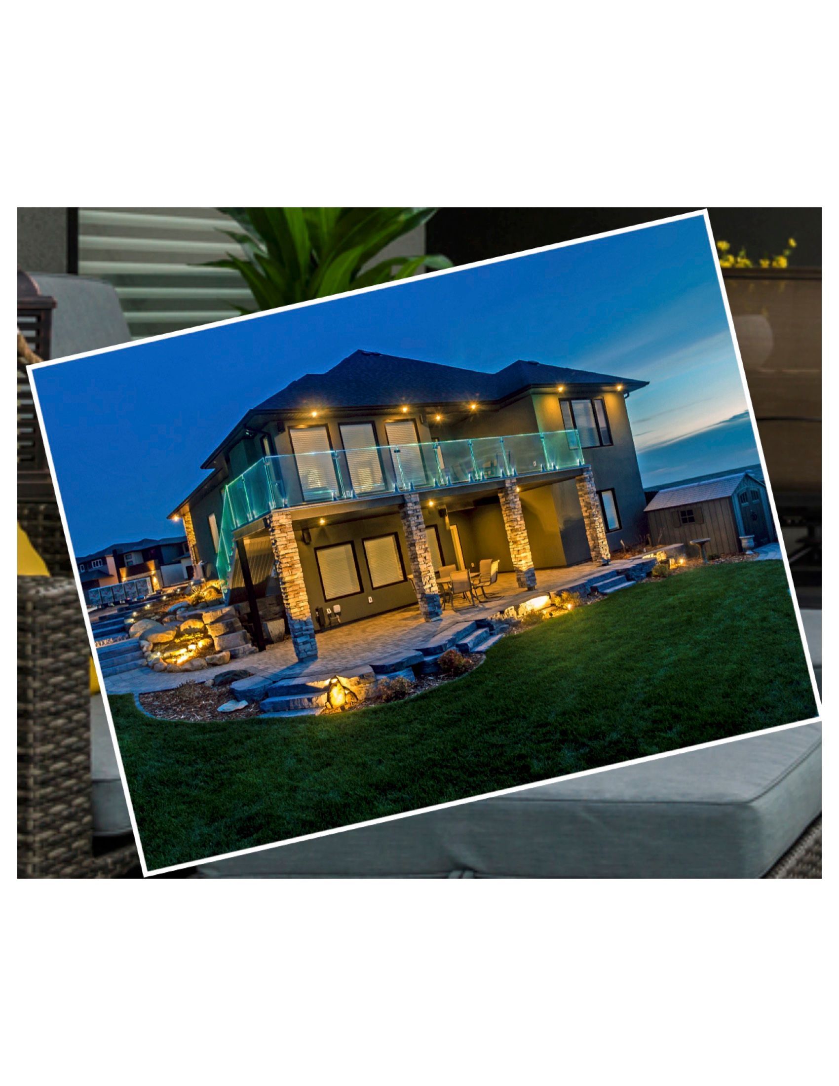 Two-story house with lights on at dusk. Stone columns, glass balcony, and landscaping.