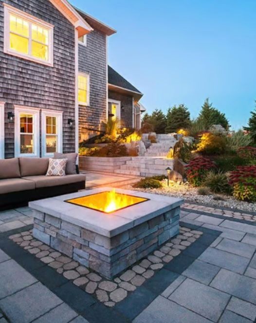 A stone fire pit with a lit flame sits in a backyard patio setting, next to a grey-shingled house.