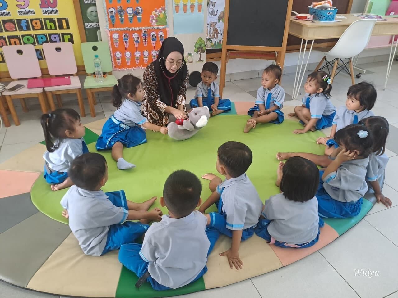 A teacher reads to children seated in a circle on a green mat in a classroom.