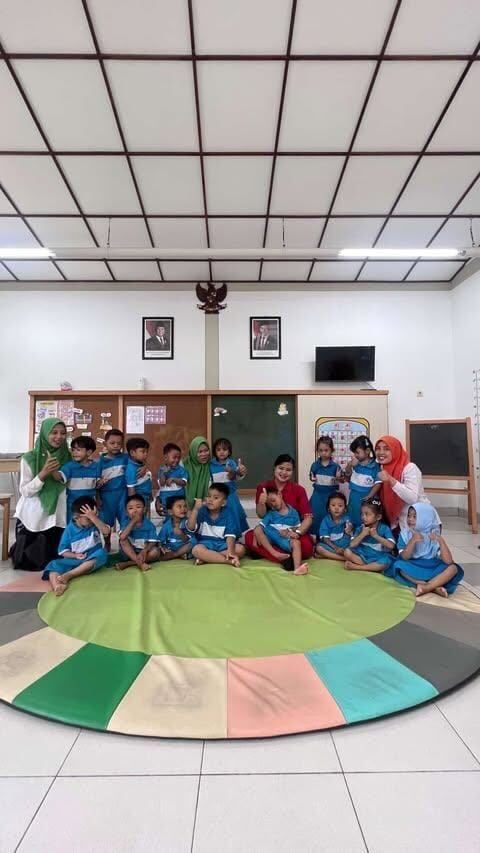 Children and adults in a classroom sitting on a colorful mat. Smiling, posing for a photo.