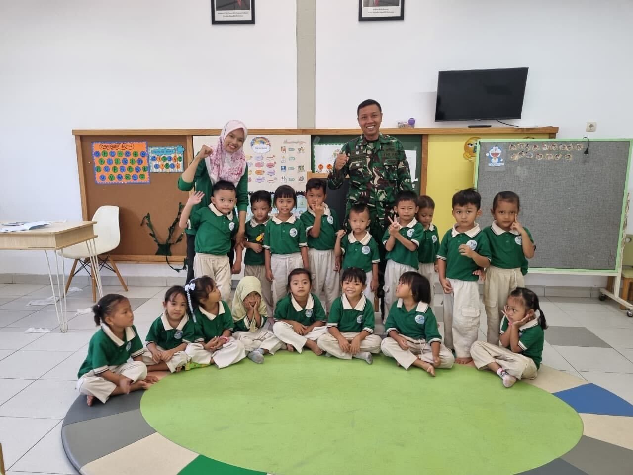 Group photo: children in green shirts with adults in a classroom, smiling.