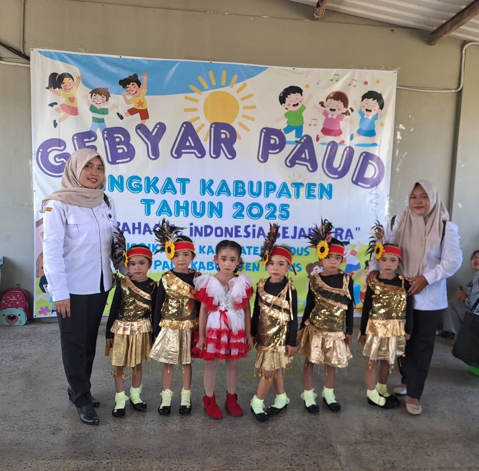 Children in costumes with teachers in front of a banner that reads “GEBYAR PAUD TINGKAT KABUPATEN TAHUN 2025”.